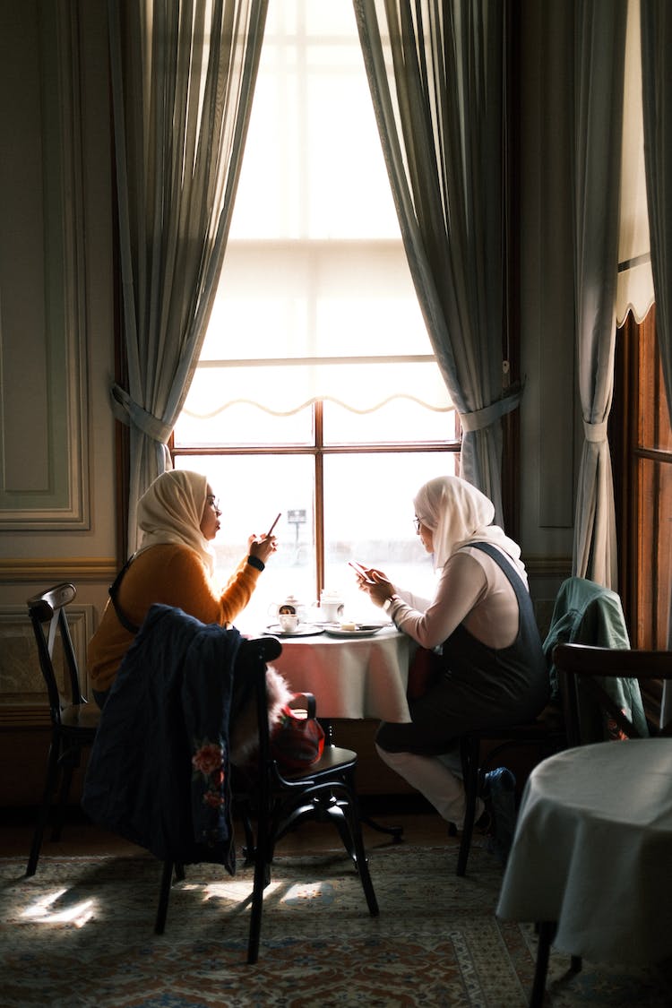 mujeres sentadas junto a una mesa debajo de una ventana y usando sus telefonos